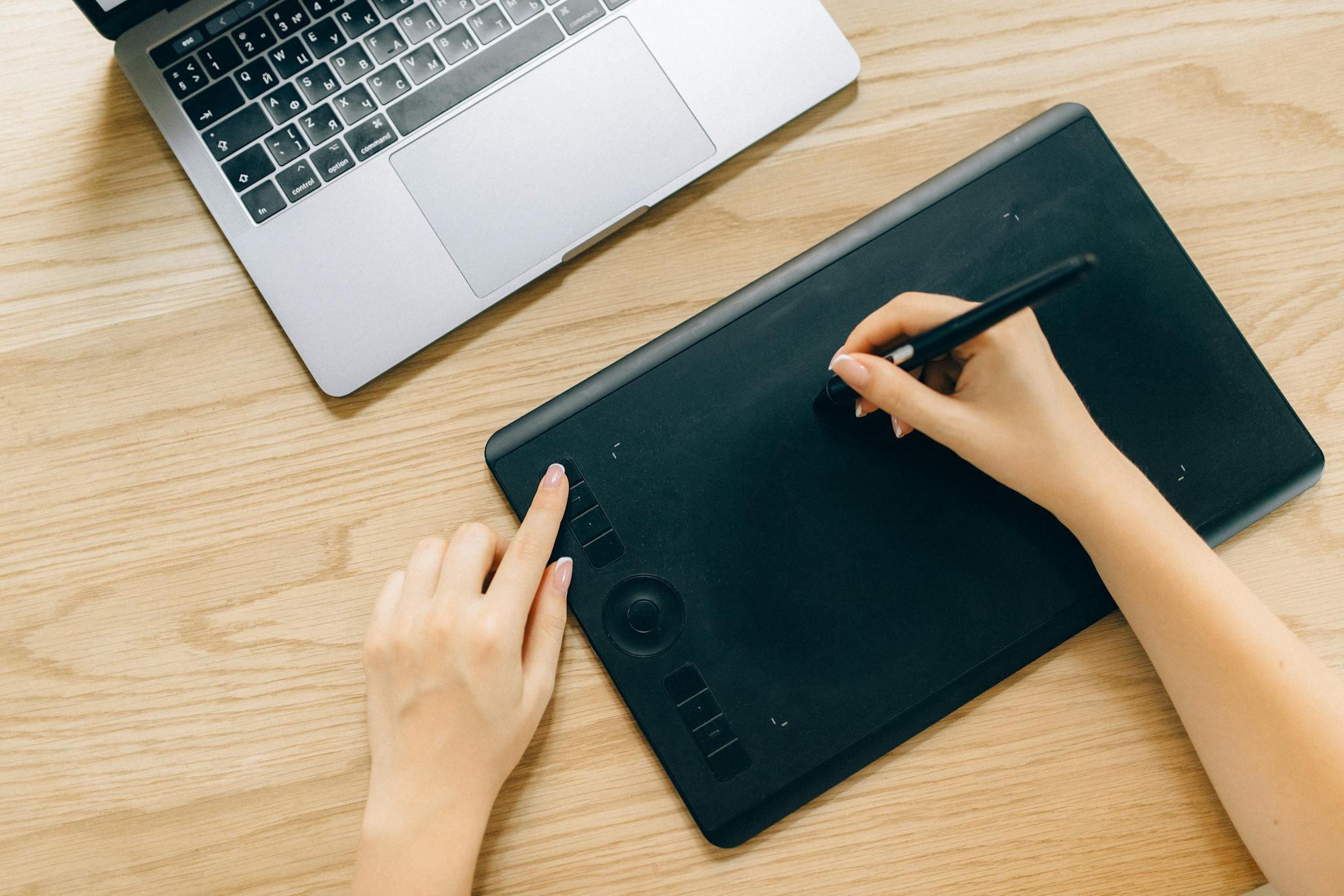 A person using a digital tablet and laptop on a wooden desk for creative work.